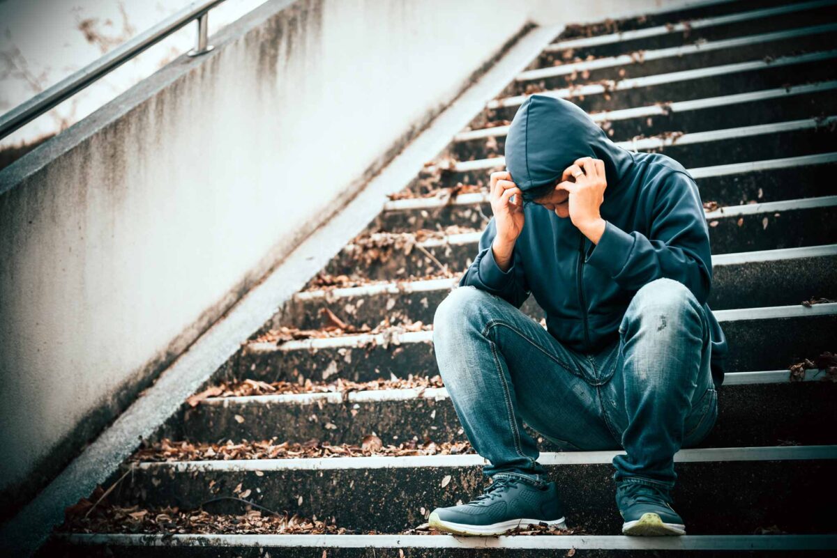 Image of a young man sitting on stairs with a hood over his head and exhibiting signs of drug use